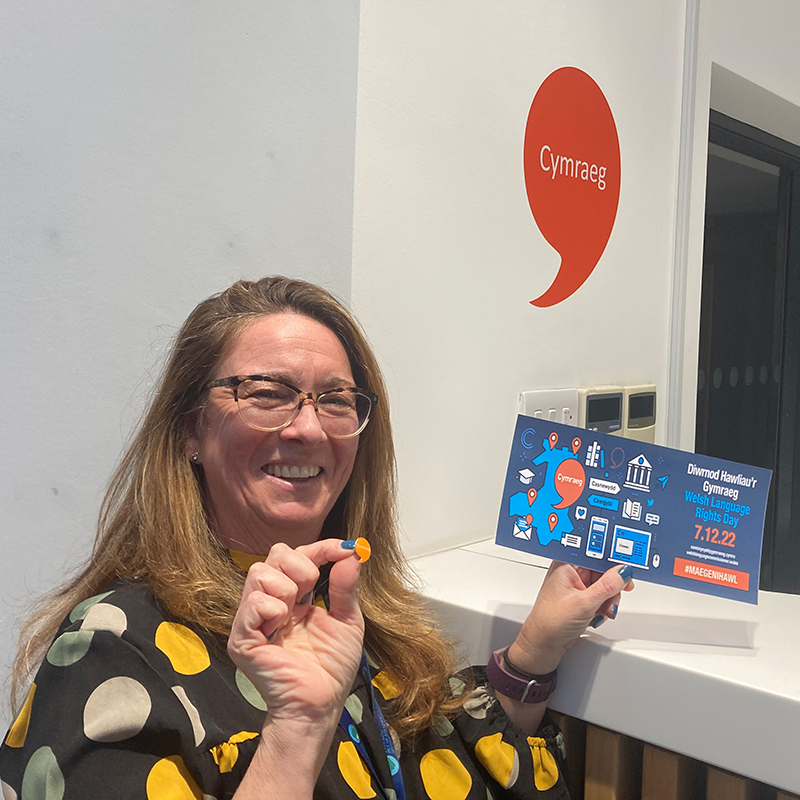 Image of an employee holding a Welsh language speaker badge and promotional flyer